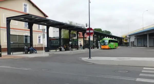 Zielony autobus FlixBus na przystanku autobusowym z wiatą i ławkami, gdzie siedzą pasażerowie.